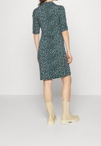 Femme portant une robe verte à fleurs arrivant au genou avec des manches jusqu'aux coudes et des bottes beige à mi-mollet, se tenant devant un fond uni.
