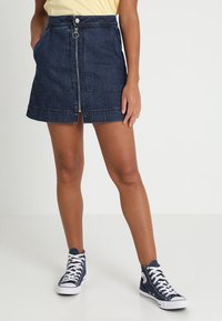 Gonna di jeans con zip frontale, colore blu scuro, due tasche laterali e orlo sopra il ginocchio, abbinata a sneakers alte navy.