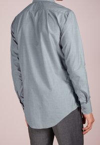 Camicia a maniche lunghe grigio chiaro con texture liscia, polsini con bottoni e orlo arrotondato, indossata sopra pantaloni grigi su sfondo rosa.