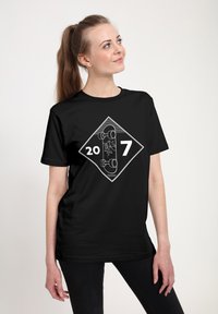 Žena s dlouhým ohonem, oblečená v černém tričku s bílým grafickým vyobrazením skateboardu a čísly "20" a "7" na diamantovém pozadí.
