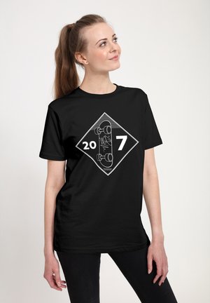 Žena s dlouhým ohonem, oblečená v černém tričku s bílým grafickým vyobrazením skateboardu a čísly "20" a "7" na diamantovém pozadí.