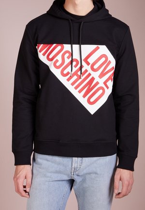 Czarna bluza z kapturem z białym ukośnym panelem i wyrazistym czerwonym napisem "LOVE MOSCHINO," noszona z jasnoniebieskimi jeansami na stojącej osobie.