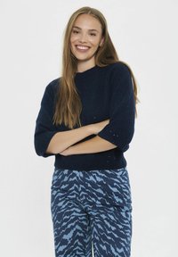 Pletený námorní sveter s krátkymi rukávmi a žebrovaným lemom, skombinovaný s modrými nohavicami so vzorom vlniek. Model stojí so prepletenými rukami.
