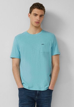 Jonge man met kort krullend haar, gekleed in een lichtblauw T-shirt met korte mouwen en borstzakje, en een donkerblauwe spijkerbroek, staand tegen een grijze muur.