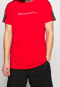 Camiseta de algodón roja con mangas cortas, escote redondo y texto cursivo blanco "Kings Will Dream". Rayas negras en las mangas.