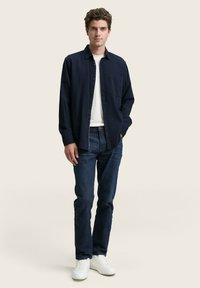 Marineblauwe overhemd met knopen over een wit T-shirt, gecombineerd met donkerblauwe jeans en witte sneakers, met een zakdetail op het shirt.