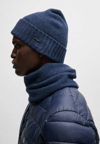 Wool knit navy beanie with ribbed cuff and matching scarf, worn over a shiny, padded navy jacket. Features a small metal logo detail.
