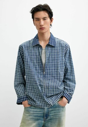 Junger Mann mit schwarzen Haaren, der ein blau-weiß kariertes Zip-up-Hemd über einem grauen Oberteil und hellblauen Jeans trägt, Hände in den Taschen, neutraler Hintergrund.