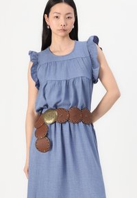 Abito in chambray blu con maniche a volant e corpino arricciato, abbinato a una cintura circolare marrone con accentuazioni in metallo e occhielli.