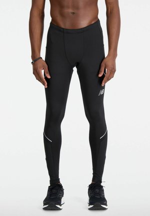 Mann in schwarzen New Balance Lauftights und schwarzen Sneakers, vor einem weißen Hintergrund stehend, mit Fokus auf Unterkörper und Beine.