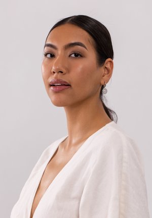 Weißes Leinenkleidungsstück mit einem tiefen V-Ausschnitt. Das Model trägt silberne, runde Ohrringe. Glatte Haut mit einem dezenten Lippenstift und zurückgestecktem Haar.