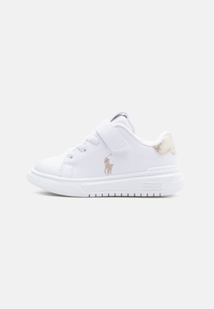 Valkoinen lasten tennari, jossa on nauhat ja tarranauha, kultainen Polo-logo sivulla ja kultainen korostus kantapäällä, litteä pohja.