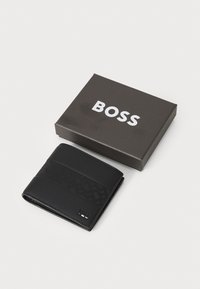 Melnas ādas maciņš ar strukturētu joslu, prezentēts tumši pelēkā kastē ar vārdu "BOSS" izceltā baltā krāsā.