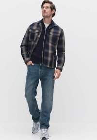 Rutig skjortjacka i marinblått och grått, med en dragkedjad marinblå stickad tröja under, kombinerat med blå jeans och grå sneakers. Två bröstfickor.