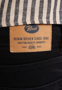 Tessuto in denim nero con etichetta in pelle color cuoio che presenta il logo "Petrol", il testo "DENIM DRIVEN SINCE 1989" e dettagli di cucitura. Tessuto a righe visibile sopra.