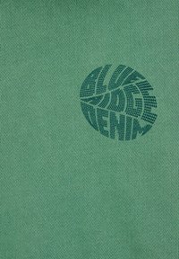 Tejido de punto verde con un patrón de hojas verde oscuro y el texto "ALIVE DENIM" centrado en la parte frontal. Textura suave.