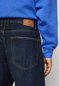 Jeans de denim oscuro con un bolsillo trasero visible, costuras amarillas en contraste y una etiqueta de cuero marrón en la cinturilla. Manga de suéter azul a la vista.