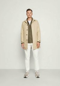 Beżowa lekka kurtka z guzikami, nałożona na oliwkową koszulę, w komplecie z białymi spodniami i beżowymi trampkami. Minimalistyczny design.