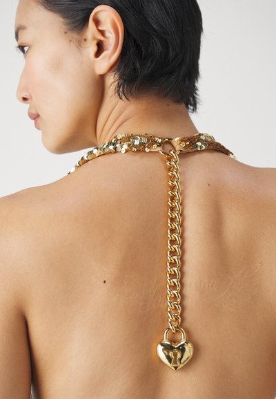 Kuldse quick-paistega krae viib paksu kuldketini, mille lõpp on südamekujulise luku vormis. Tekstuur on läikiv ja metalne, kontrastides naha tooniga.