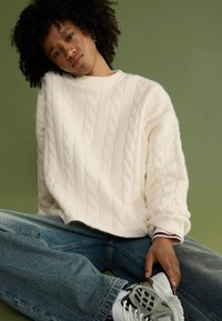 Översize krämfärgad kabelstickad tröja med ribbat halsband, parat med lösa denimjeans och grå sneakers med mesh-detaljer.