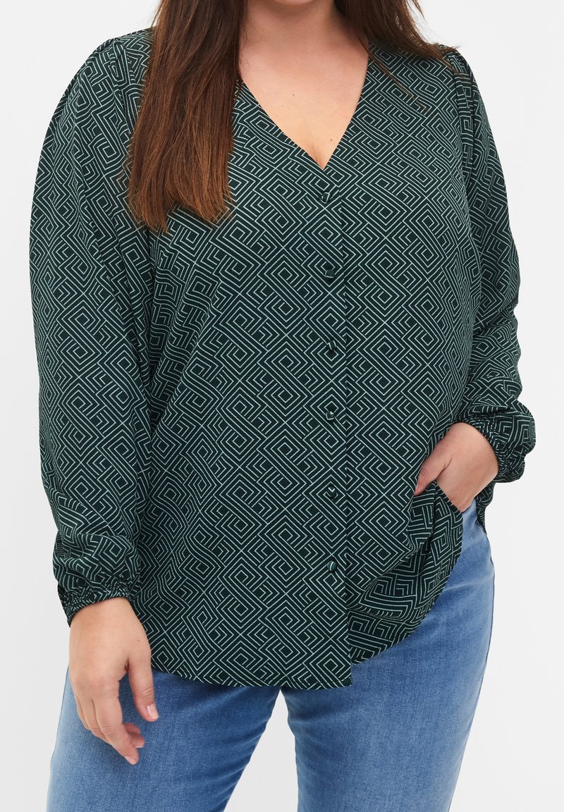 Femme portant un chemisier vert à manches longues avec un motif géométrique blanc et un jean bleu, une main dans la poche, debout devant un fond uni.