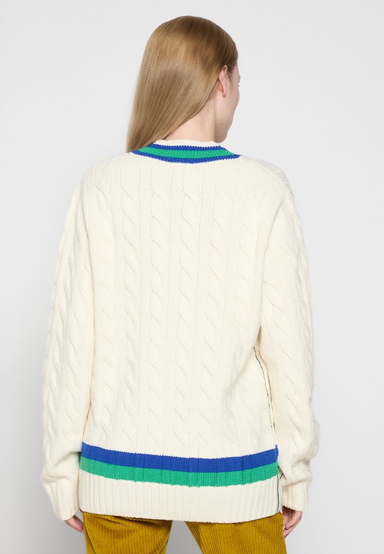 Maglione crema a treccia con colletto e orlo a strisce blu e verde a coste. Presenta una vestibilità rilassata e spacchi laterali per facilitare i movimenti.