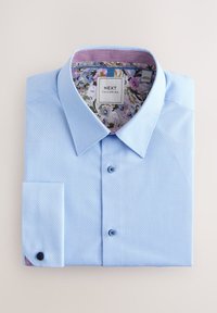 Lyseblå skjorte med knapper, tekstureret stof, klassisk krave og blomstret inderfor. Har mørke knapper og enkelt brystlomme.