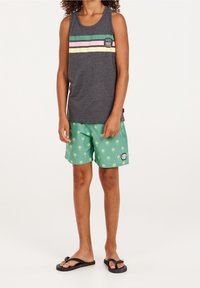 Bambino che indossa una maglietta senza maniche grigio scuro con strisce colorate e pantaloni corti verdi con stampa di palme e infradito nere, in piedi su uno sfondo bianco.