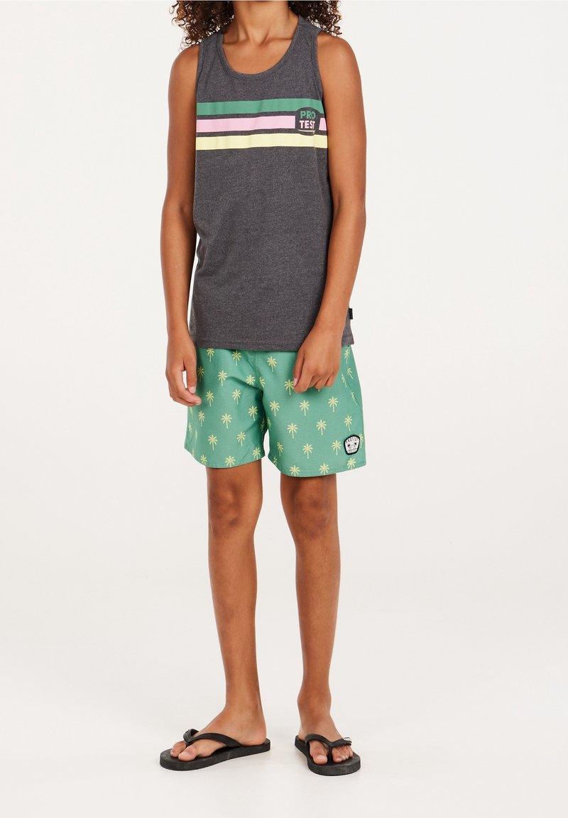 Bambino che indossa una maglietta senza maniche grigio scuro con strisce colorate e pantaloni corti verdi con stampa di palme e infradito nere, in piedi su uno sfondo bianco.