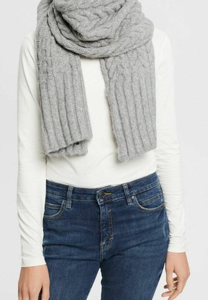 Personne portant une grosse écharpe en maille torsadée gris clair, une chemise blanche à manches longues et un jean bleu avec les poches avant visibles.