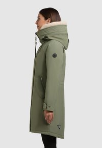 Olivovozelená parka s kapucňou vystlanou kožušinou, zipsovými vreckami a nášivkou s logom. Má priamy strih a nastaviteľné manžety.