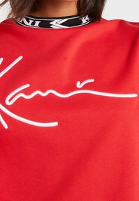 Sudadera roja con un diseño de firma bordada en blanco. Cuello a rayas en blanco y negro con detalles estampados. Tejido suave y texturizado.