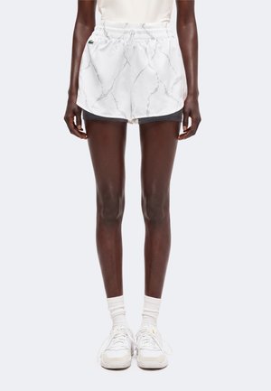 Persoon draagt witte sportshorts met patroon over zwarte compressie shorts, witte sokken en witte sneakers, staand tegen een effen achtergrond.