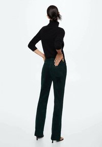 Musta poolapaita yhdistettynä korkeavyötäröisiin tummanvihreisiin corduroy-housuihin. Malli seisoo kädet lanteilla, esitellen asua takaa.