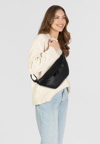 Borsa crossbody nera in pelle con forma curva, texture liscia e tracolla regolabile, indossata di lato da una persona in abbigliamento chiaro.