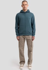 Felpa con zip di colore verde acqua, con lacci, due tasche frontali e polsini a costine. Abbinata a pantaloni cargo beige e sneakers color crema. Design semplice.