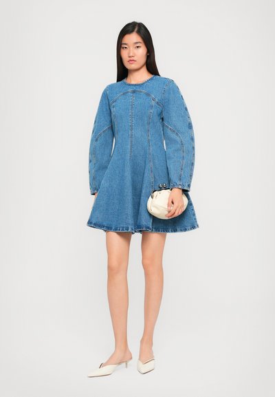 Džínová sukňa s rozšíreným A-čkovým strihom, dlhými volánovými rukávmi a kontrastným prešitím. Skombinovaná s malou bielou kabelkou a krémovými šľapkami.