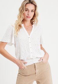 White short-sleeve blouse with eyelet embroidery, v-neckline, and button detail, featuring a gathered waist and relaxed fit. Paired with beige pants.
