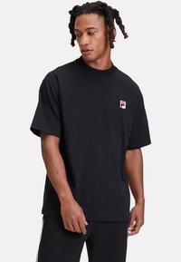 Svart t-shirt med kort ärm i bomull, med en liten broderad logotyp i röd och blå nyans på bröstet och en rund halsringning.