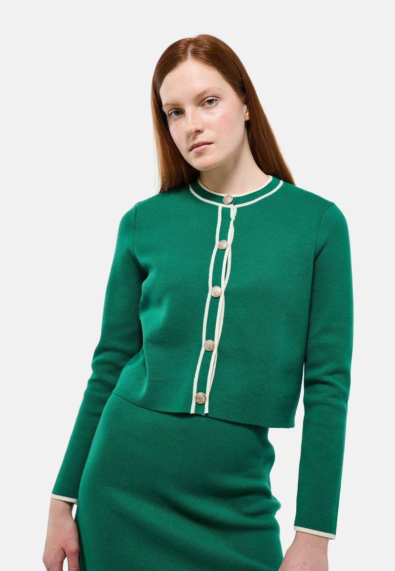 Pull vert avec un col rond, des bordures blanches et cinq boutons décoratifs sur le devant. Fabriqué en tissu en tricot texturé.