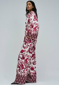 Mujer llevando un vestido largo blanco con grandes patrones florales rojos y mangas anchas, de perfil contra un fondo claro y liso.