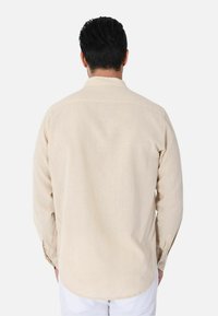Camicia beige a maniche lunghe realizzata in tessuto leggero, con colletto alla francese e polsini con un bottone, dotata di orlo curvo.