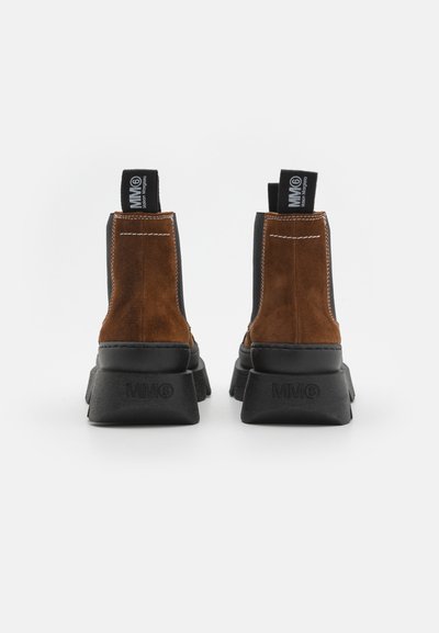 Botas de tobillo de ante marrón con una suela de goma negra, que presentan costuras en contraste y lengüetas en la parte posterior para facilitar el uso.