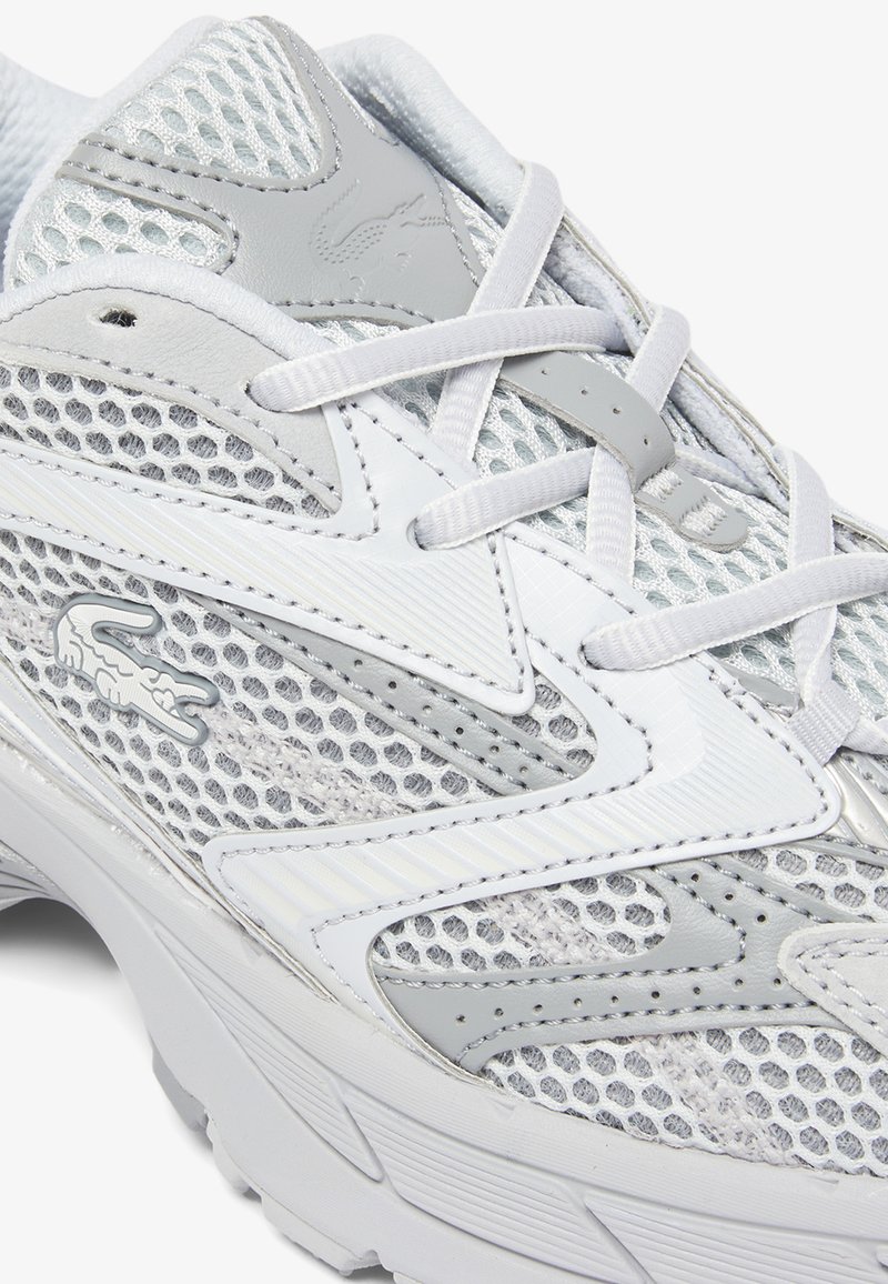 Scarpa sportiva bianca e grigia con tessuto in mesh, cuciture dettagliate, lacci annodati e logo di coccodrillo sul lato.