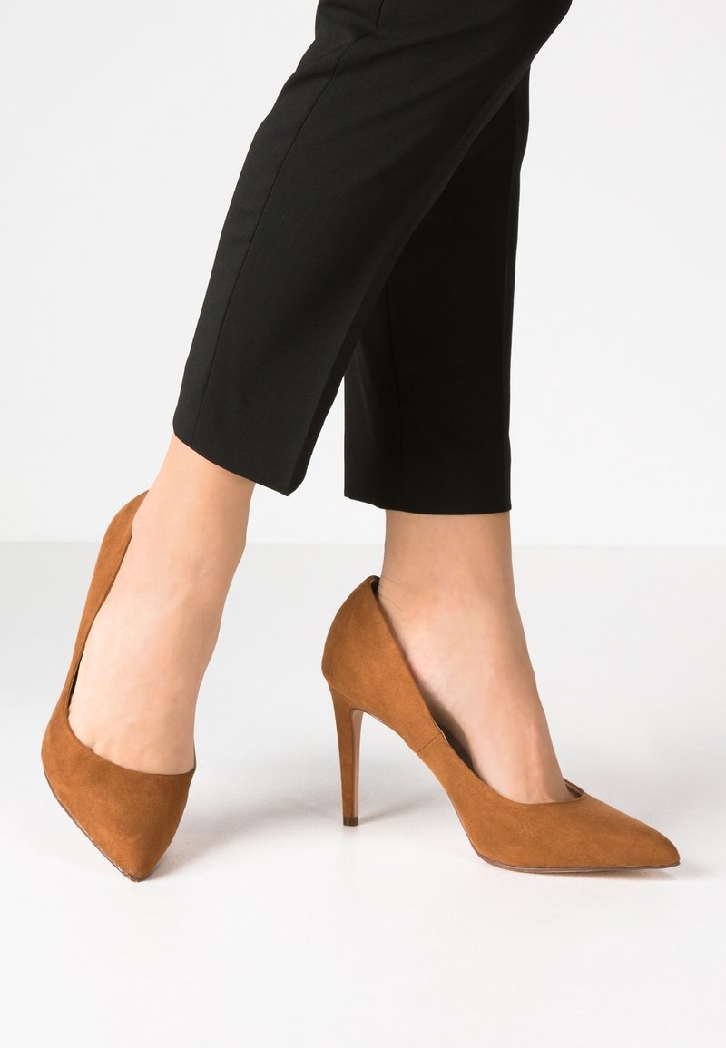 Jambes portant un pantalon court noir et des chaussures à talons hauts en daim marron à bout pointu sur une surface blanche.