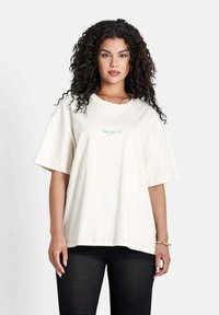 T-shirt beige oversize à manches courtes, avec un logo en texte bleu sur le devant. Le tissu semble doux, avec une coupe décontractée. Porté avec un pantalon en mesh noir.