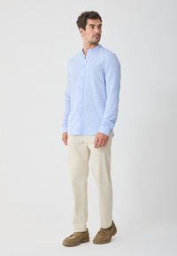 Light blue long-sleeve shirt with a mandarin collar, button closure, and subtle texture, paired with cream-colored trousers and brown shoes.