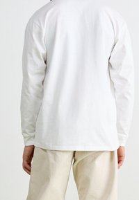 Persoon in een effen wit lange mouwen shirt en lichtbeige broek, staand met de rug naar de kijker tegen een effen achtergrond.