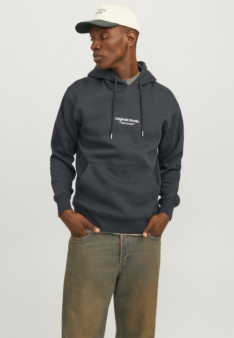 Mørk grå hettegenser med lomme foran og snor, med et hvitt logo-print. Båret med falmede khaki jeans og en lys lue.