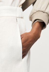 Χέρι με σκούρο δέρμα που ακουμπάει μέσα στην τσέπη λευκού παντελονιού, φοράει μπεζ σακάκι με ριμπ μανσέτα σε ανοιχτό φόντο.